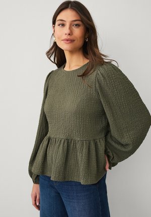 Kvinde iført en olivengrøn tekstureret bluse med lange ærmer, puffede ærmer og indsnævret talje, kombineret med blå jeans, foran en ensfarvet baggrund.