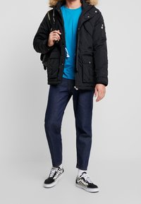 Svart puffjacka med fuskpäls på huven, kombinerad med en turkos skjorta, mörka jeans och svartvita rutiga sneakers.