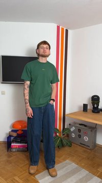 Groene T-shirt met korte mouwen, blauwe wijde jeans, bruine laarzen. Tatoeages zichtbaar op de armen. Achtergrond bevat kleurrijke muurstrepen en meubels.