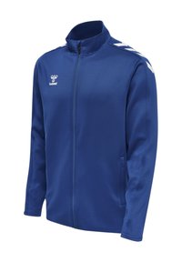 Chaqueta deportiva azul con cremallera completa en el frente, logo blanco de hummel en el pecho y rayas blancas en forma de chevrón en los hombros.