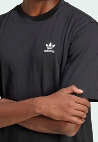 Donkerhuidige persoon in een zwart Adidas T-shirt met een wit logo, met gekruiste armen en één hand die de tegenoverliggende bovenarm vasthoudt.