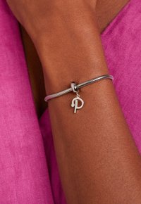 Zilveren armband met een flexibele ontwerpelement, voorzien van een ronde charm die gegraveerd is met een gestileerde "P" en een kleine strass accent.
