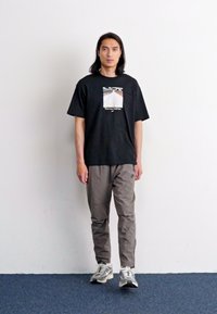 Tricou negru cu mânecă scurtă și un grafic pe piept, asortat cu pantaloni ușor gri, ajustați și pantofi sport argintii.