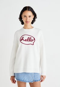 Pull en blanc avec un design de bulle de parole "bonjour" en maroon, avec une coupe décontractée et des poign wrists côtelés, associé à une jupe en denim.