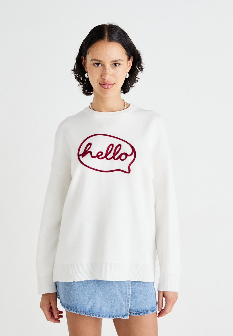 Pull en blanc avec un design de bulle de parole "bonjour" en maroon, avec une coupe décontractée et des poign wrists côtelés, associé à une jupe en denim.