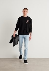 Hanorac negru cu detalii grafice, blugi albaștri deschiși și adidași negri. Modelul ține o geacă neagră, stând în fața unui fundal neutru.