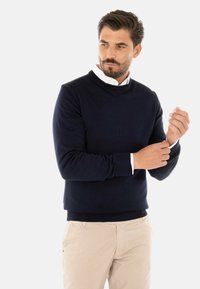 Maglione girocollo navy realizzato in morbido tessuto, con maniche lunghe e polsini a costine, indossato sopra una camicia bianca con colletto.