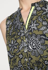 Vestido sin mangas de tela color verde oliva con un patrón floral en blanco y negro, que presenta un cierre de botones en el frente y un acento en amarillo neón a lo largo del escote.