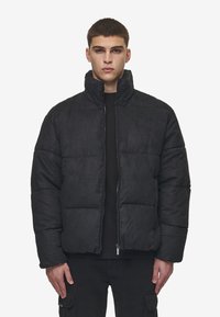 Svart pufferjacka i mjukt material, med hög krage, dragkedja och vadderade sektioner för isolering. Minimal märkning synlig.