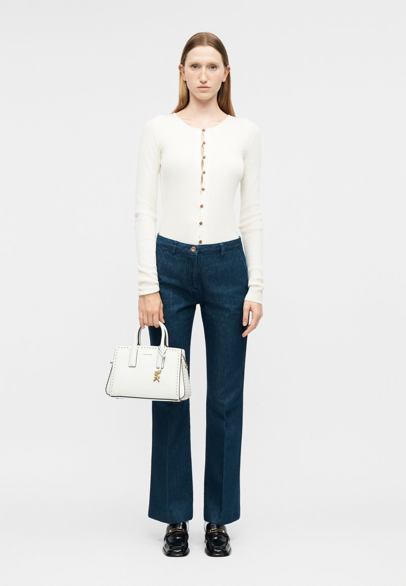 Γυναίκα που φοράει ένα λευκό πλεκτό ζακέτα με κουμπιά, σκούρα μπλε στενά τζιν κανονικού μήκους, μαύρα μοκασίνια, κρατώντας μια λευκή δομημένη τσάντα χειρός με το αριστερό της χέρι.