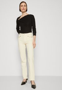 Top negro de manga larga con cuello asimétrico y acento de joyas, combinado con pantalones color crema que presentan detalles de metal dorado.