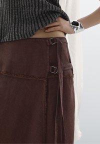 Lähellä otettu kuva henkilöstä, joka on pukeutunut harmaaseen ribbivillapaitaan ja ruskeaan kietaisuhameeseen, jossa on metalliset soljeyksityiskohdat. Henkilön käsi on asetettu vyötärölle, ja hänellä on hopeaketju ranteessaan.