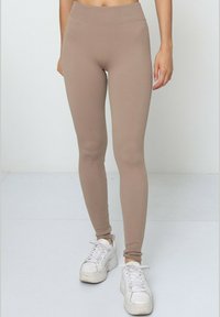 Leggings beige à côtes avec une taille haute, un design fuselé et une texture douce, associés à des baskets blanches sur un fond clair.