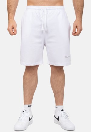 Mann trägt weiße Shorts mit Kordelzug, weiße Puma-Socken und weiße Nike-Sneaker, steht vor einem einfarbig hellen Hintergrund.