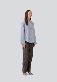 Stribet skjorte med knapper, blå og hvid, med oversized ærmer. Båret med brune læderbukser og snøre sandaler. Glat tekstur.