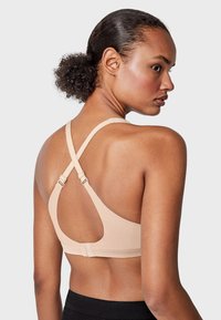 Sujetador beige hecho de un material suave y elástico, con tirantes en forma de cruz en la parte posterior y herrajes ajustables para un mejor ajuste. Diseño minimalista.
