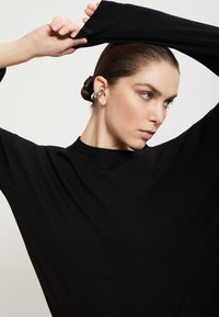 Maglietta nera a maniche lunghe con vestibilità ampia e polsini a costine. Il modello presenta un design semplice e minimalista e un accessorio per orecchio.