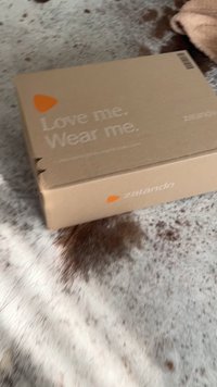 Tekturowe pudełko wysyłkowe o jasnobrązowej powierzchni. Tekst brzmi "Kochaj mnie. Noś mnie." i zawiera pomarańczowe logo Zalando.