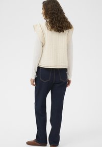 Prošívaná béžová vesta s nařasenými švy na ramenou, kombinovaná s tmavými džíny z denimu, které mají volný střih a jemné prošívání na kapsách.