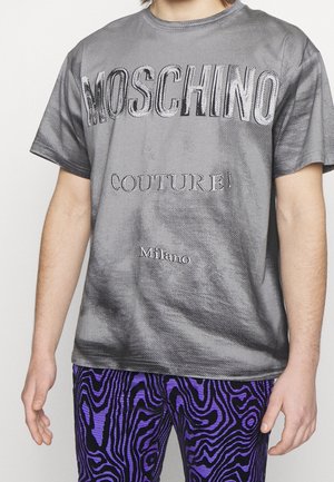 Person wearing a gray Moschino t-shirt with "Couture!" and "Milano" text, paired with purple and black patterned pants.