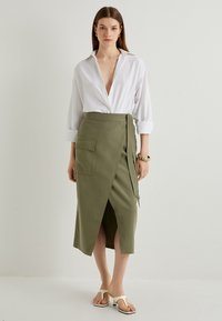 Chemise blanche à manches longues associée à une jupe portefeuille verte dotée d'une poche latérale, d'une fente haute et d'un détail de nouage assorti. Sandales blanches plates.