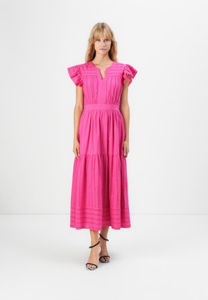 Woman standing against white background wearing a bright pink midi dress with flutter sleeves and strappy black and white heels.