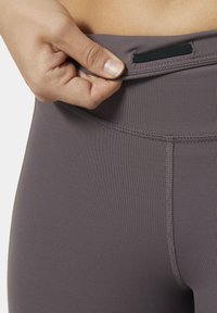 Grijze sportleggingen met een gladde textuur, een elastische tailleband en gestikte details. Hand grijpt de tailleband voor aanpassing.