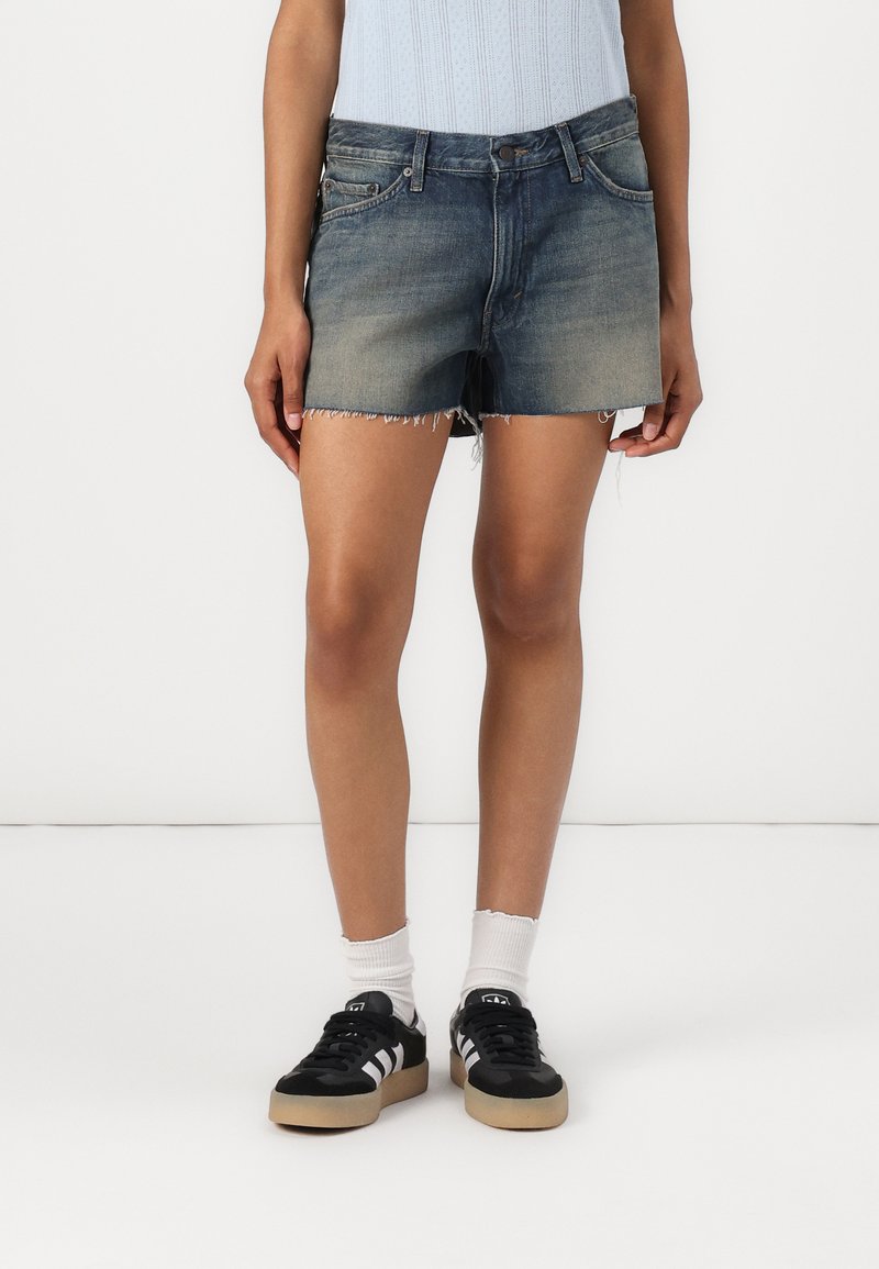 Denim short in een vervaagde blauwe kleur, met rafelige uiteinden, een vijf-pocket ontwerp en een aansluitende pasvorm. Gecombineerd met zwarte sneakers.