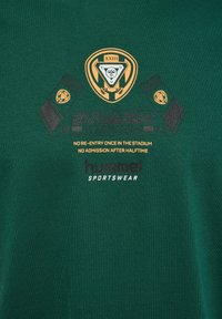 Ciemnozielona koszulka bawełniana z graficznym wzorem z czarnymi i złotymi akcentami, tekstem i logo, umieszczona na środku w okolicy klatki piersiowej.