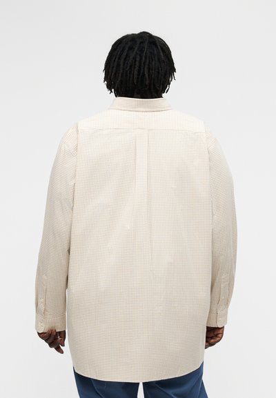 Osoba s tamnim dreadlocksima koja nosi bež košulju na pruge s dugim rukavima i plave hlače, prikazana sa stražnje strane na jednobojnoj pozadini.