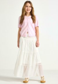 Pink kortærmet cardigan med knapper, parret med en hvid, lagdelt maxi-nederdel. Båret med beige afslappede sneakers. Enkle, bløde teksturer.