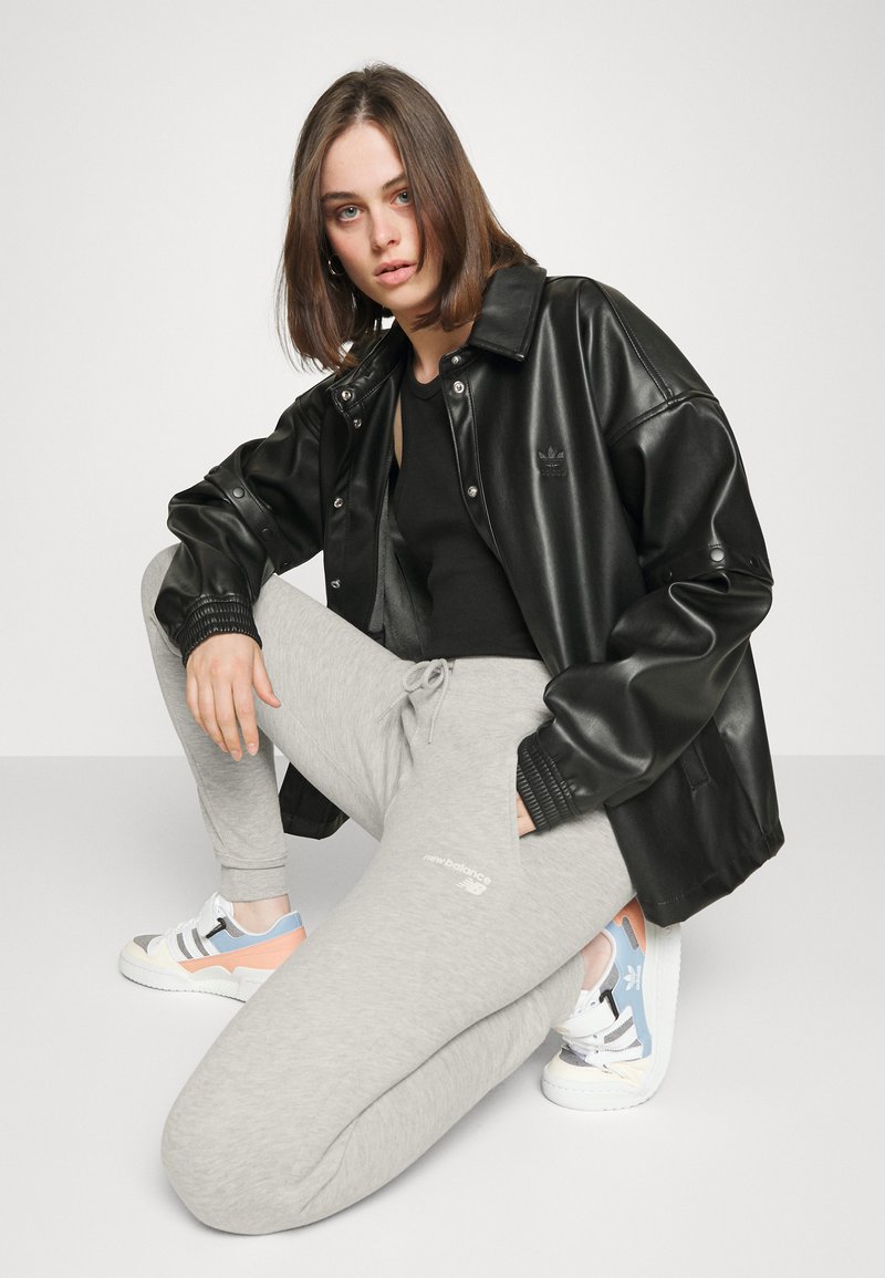 Chaqueta de cuero sintético negra, pantalones de chándal grises y zapatillas blancas con acentos en tonos pastel. Modelo de rodillas, mostrando claramente los detalles de la ropa.