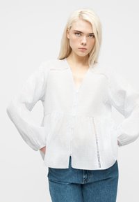 Νεαρό άτομο με μακριά ξανθά μαλλιά, φορώντας λευκό πουκάμισο με κουμπιά και διακριτικές κάθετες ρίγες και μπλε τζιν, με τα χέρια στις τσέπες.