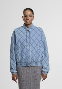 Gai zila džinsa jaka ar dimanta rakstu, snap pogām, stāvošu apkakli un elastīgām piedurknēm. Valkāta virs pelēkā ribota svārka.