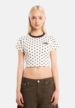 Giovane donna con capelli biondi lisci indossa un top corto bianco con pois neri e scritta marrone, abbinato a pantaloni marrone scuro.