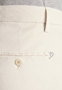Gros plan sur un pantalon couleur crème avec une poche arrière boutonnée et un petit logo métallique près de la poche.