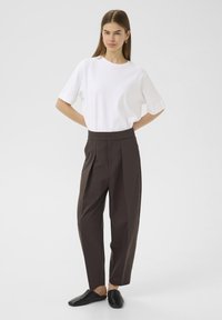 T-shirt bianco a maniche corte abbinato a pantaloni scuri marroni a vita alta con pince, indossato con scarpe nere slip-on.