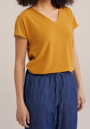 Kobieta w musztardowożółtym teksturowanym topie z krótkim rękawem i dekoltem w serek, włożonym do luźnych niebieskich spodni z wiązaniem.