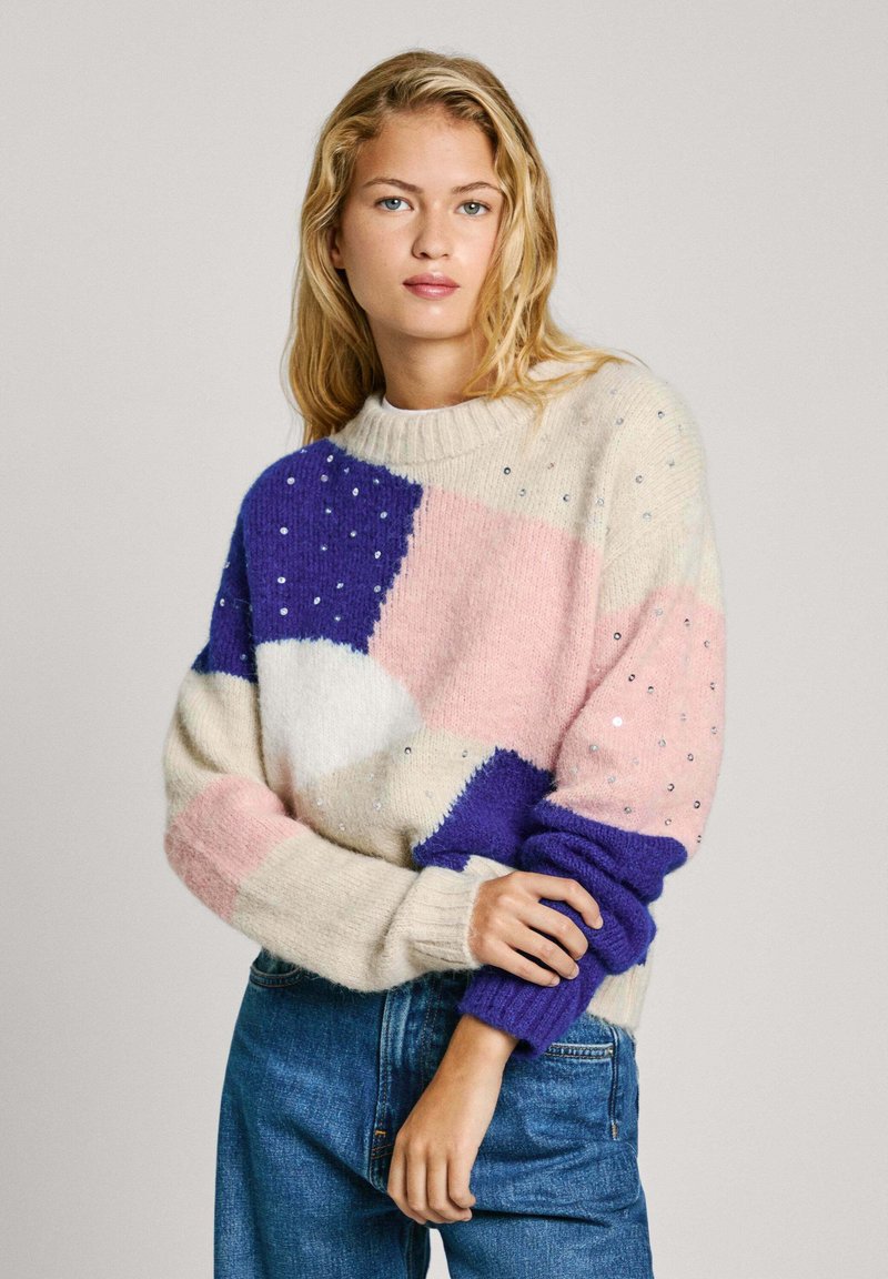 Pull en tricot avec un motif géométrique en violet, rose et crème agrémenté de détails. Poignets côtelés en bleu. Coupe décontractée.