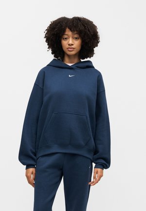 Vrouw met krullend haar die een marineblauwe Nike hoodie en bijpassende joggingbroek draagt, staand tegen een effen lichte achtergrond.