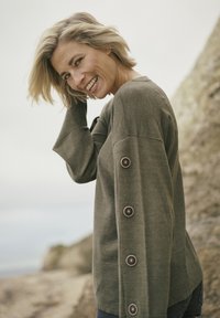 Pull olive en vert foncé avec des manches longues, accents de boutons sur le côté et coupe décontractée. Texture douce et design minimaliste.