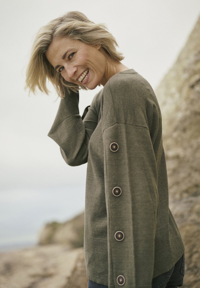 Pull olive en vert foncé avec des manches longues, accents de boutons sur le côté et coupe décontractée. Texture douce et design minimaliste.