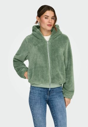 Kvinne iført en lys grønn, myk hettejakke med glidelås og ribbede mansjetter, kombinert med blå jeans, står med en hånd i lommen.