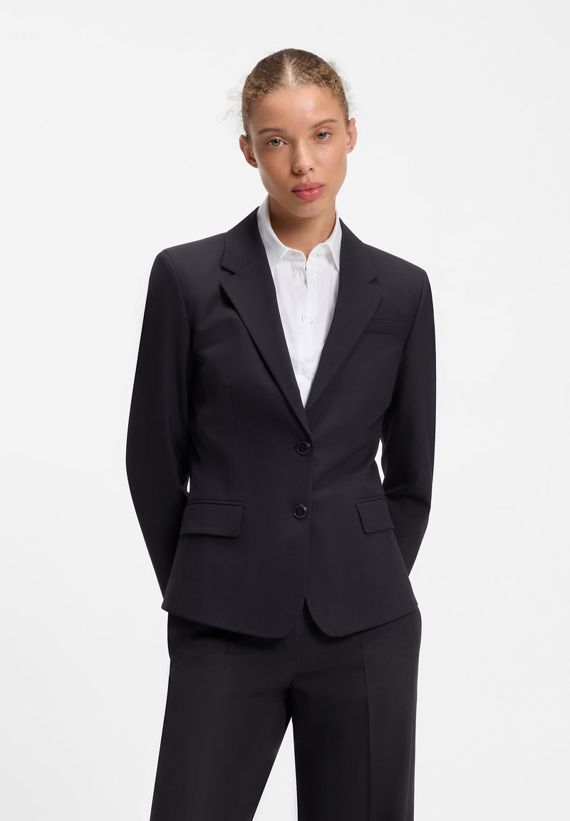 Femme aux cheveux châtain clair attachés vers l’arrière, portant une veste de costume noire ajustée et un pantalon assorti sur une chemise blanche boutonnée.
