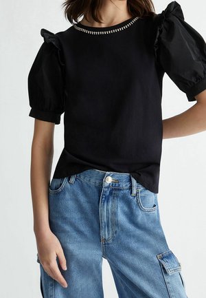 Czarny top z krótkimi, bufkami na rękawach i ozdobnym dekoltem, zestawiony z jasnoniebieskimi jeansami cargo z widocznymi kieszeniami i szwami.