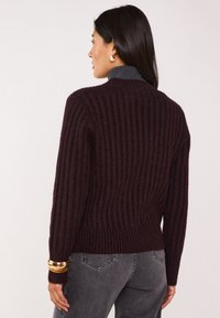 Maglione bordeaux a costine con collo alto, realizzato in tessuto lavorato a maglia con texture e design aderente. Bracciale dorato visibile al polso.