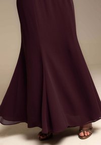 Larga falda oscura morada con pliegues cerca del dobladillo, combinada con sandalias de bronce con tiras y punta descubierta sobre un suelo beige.