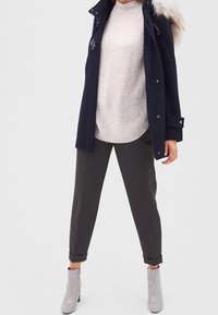 Person wearing navy fur-hooded coat, light gray sweater, dark gray cuffed pants, and shiny light gray ankle boots standing with legs apart.