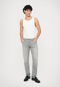 Jeans cinza claro com um corte clássico, apresentando um design de cinco bolsos e costura em contraste, combinados com uma t-shirt branca sem mangas e sapatos pretos.