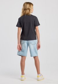 Kurze ärmeln, dunkelgraues Baumwoll-T-Shirt, kombiniert mit hellblauen Jeansshorts. Weiße Sneaker mit gelben Akzenten, von hinten gesehen.